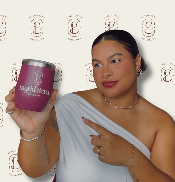 Ingrid Sena sorrindo e apontando para um copo com a marca dela, diante do painel da marca