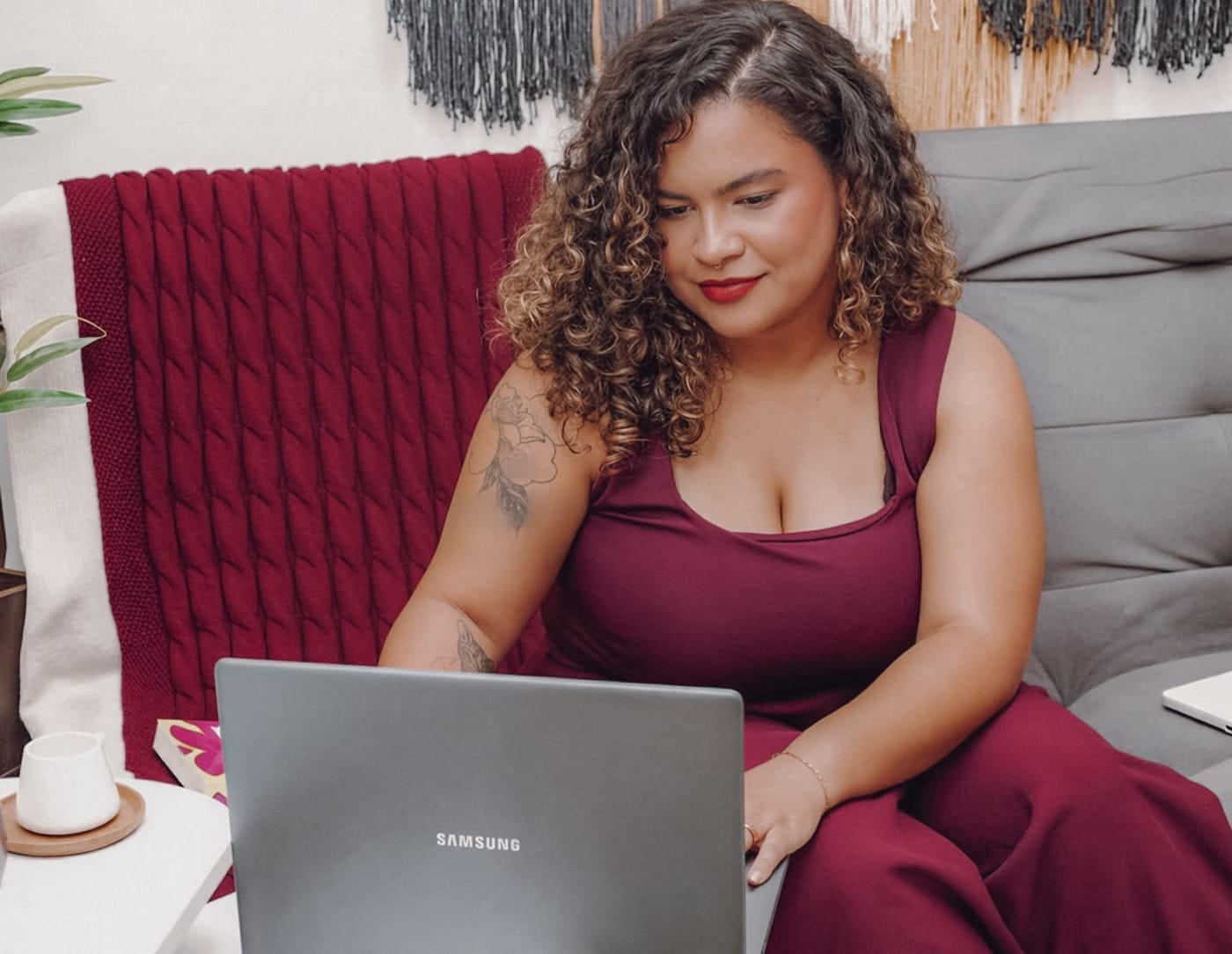 Ingrid Sena sentada no sofá, atendendo pelo notebook, com uma manta bordô ao fundo