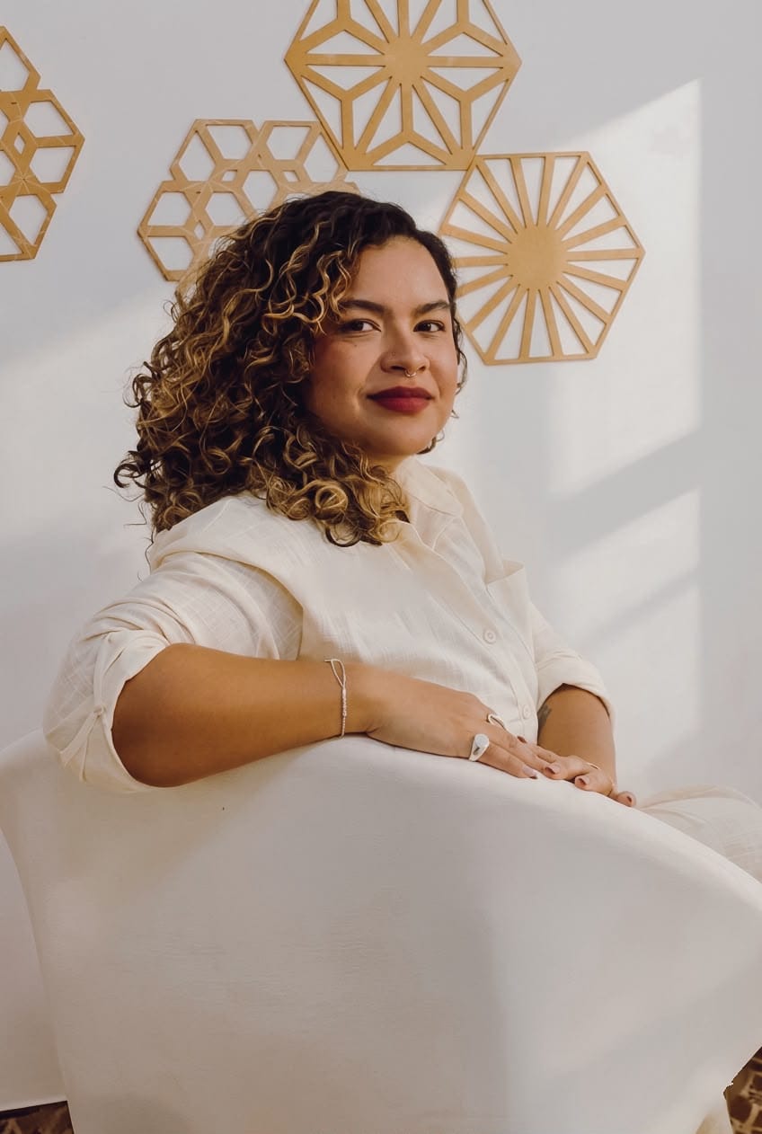 Ingrid Sena sentada em uma poltrona clara, de camisa off-white, diante de uma parede com peças geométricas douradas